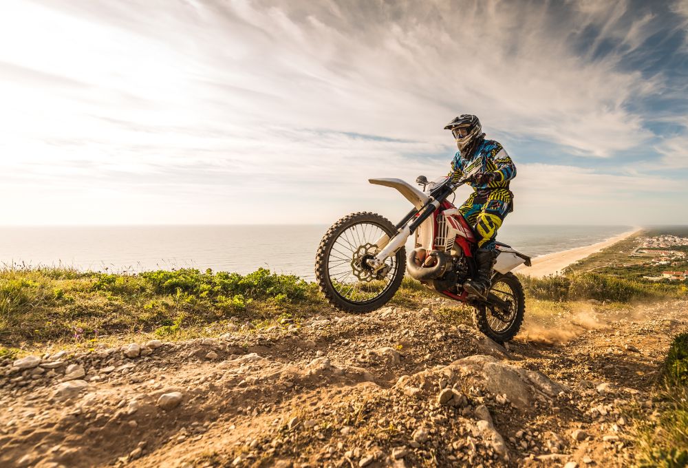 A man on a motorcycle rides a dirt road on the back tire along the coastline
